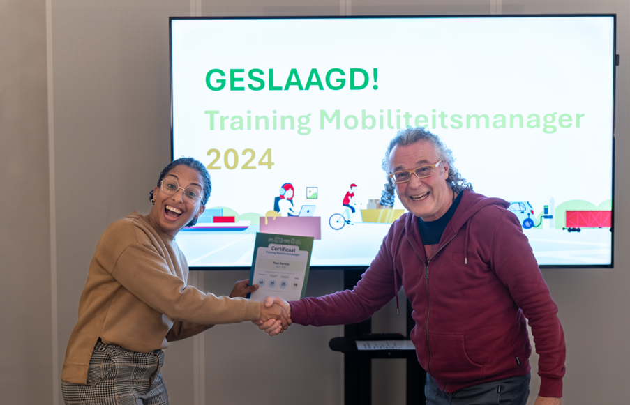 Pavi Fermin ontvangt het certificiaat van de training tot mobiliteitsmanager uit handen van Rob Schaap. Ze staan voor een wit scherm met daarop Geslaagd