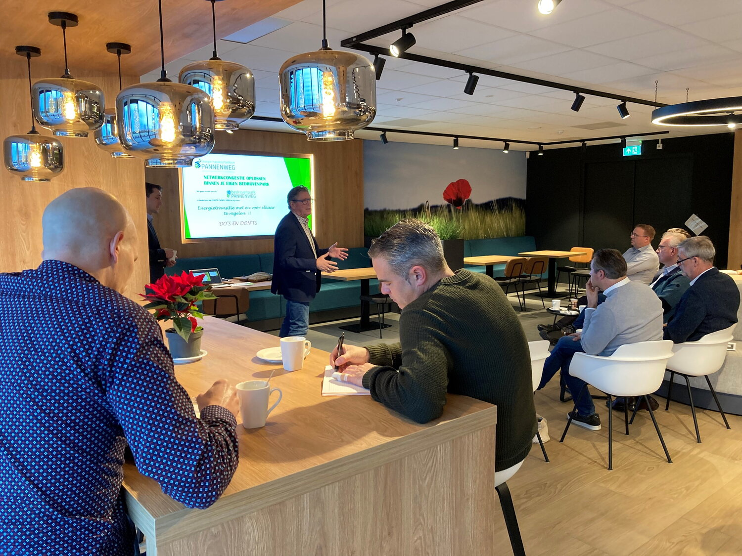 Maastrichtse ondernemers op bezoek op bedrijventerrein Pannenweg in Nederweert. Waar ze een presentatie krijgen over het verduurzamen van bedrijventerreinen. Op de foto zie je een man die presenteert terwijl zes mannen zitten op een stoel of hoge kruk terwijl ze luisteren naar de presentatie die gepresenteerd wordt op een groot scherm. 