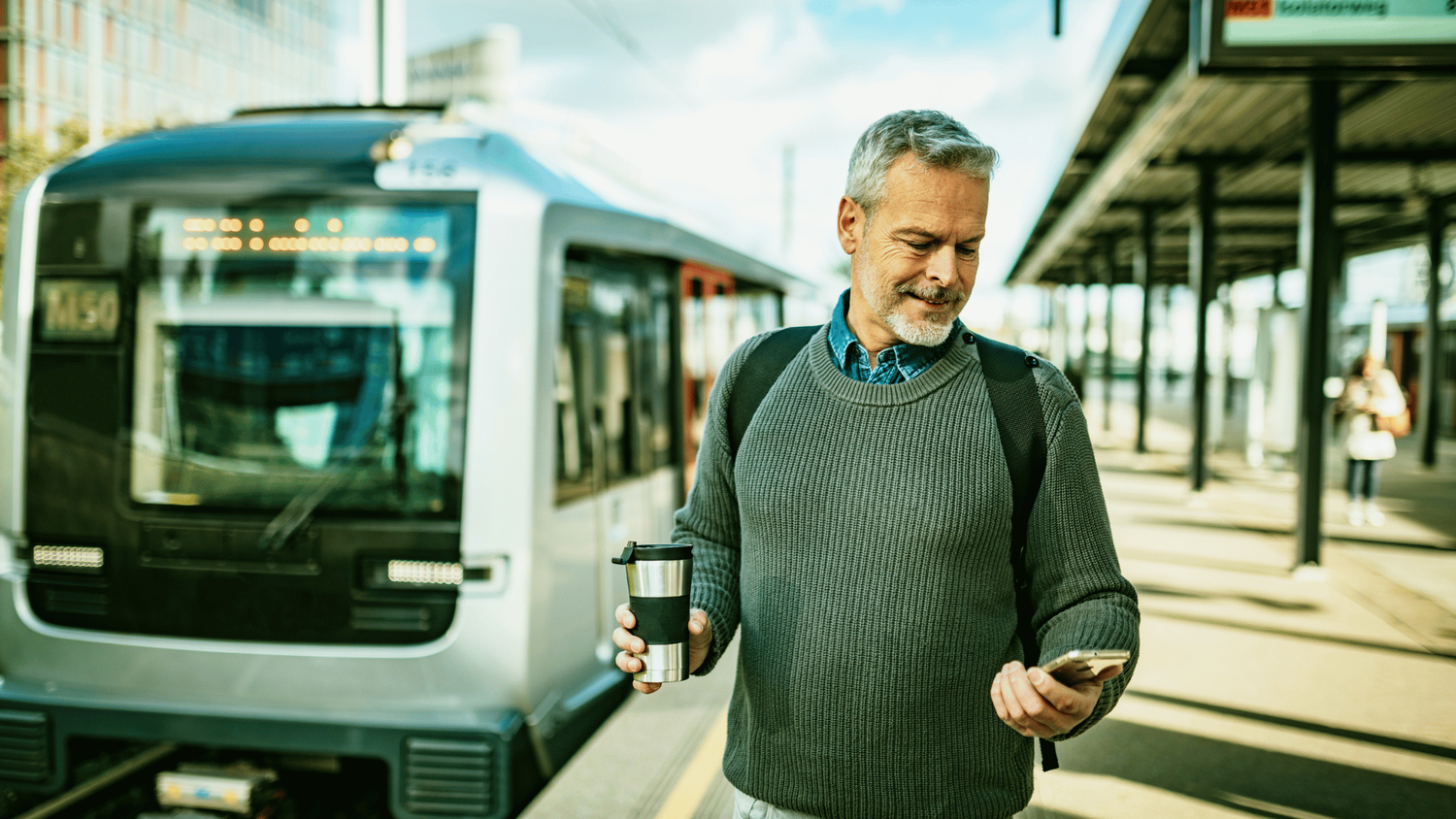 Man kijkt op de telefoon met op de achtergrond de trein. Hij loopt op het perron.