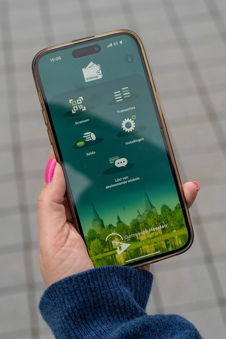 Een vrouwenhand houdt een telefoon vast met daarop het beginscherm van de Vitknip-app gemeente Heerlen